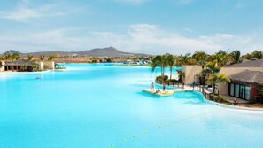 Una piscina grande y cristalina de color azul con un muelle, palmeras y edificios de un complejo turístico.