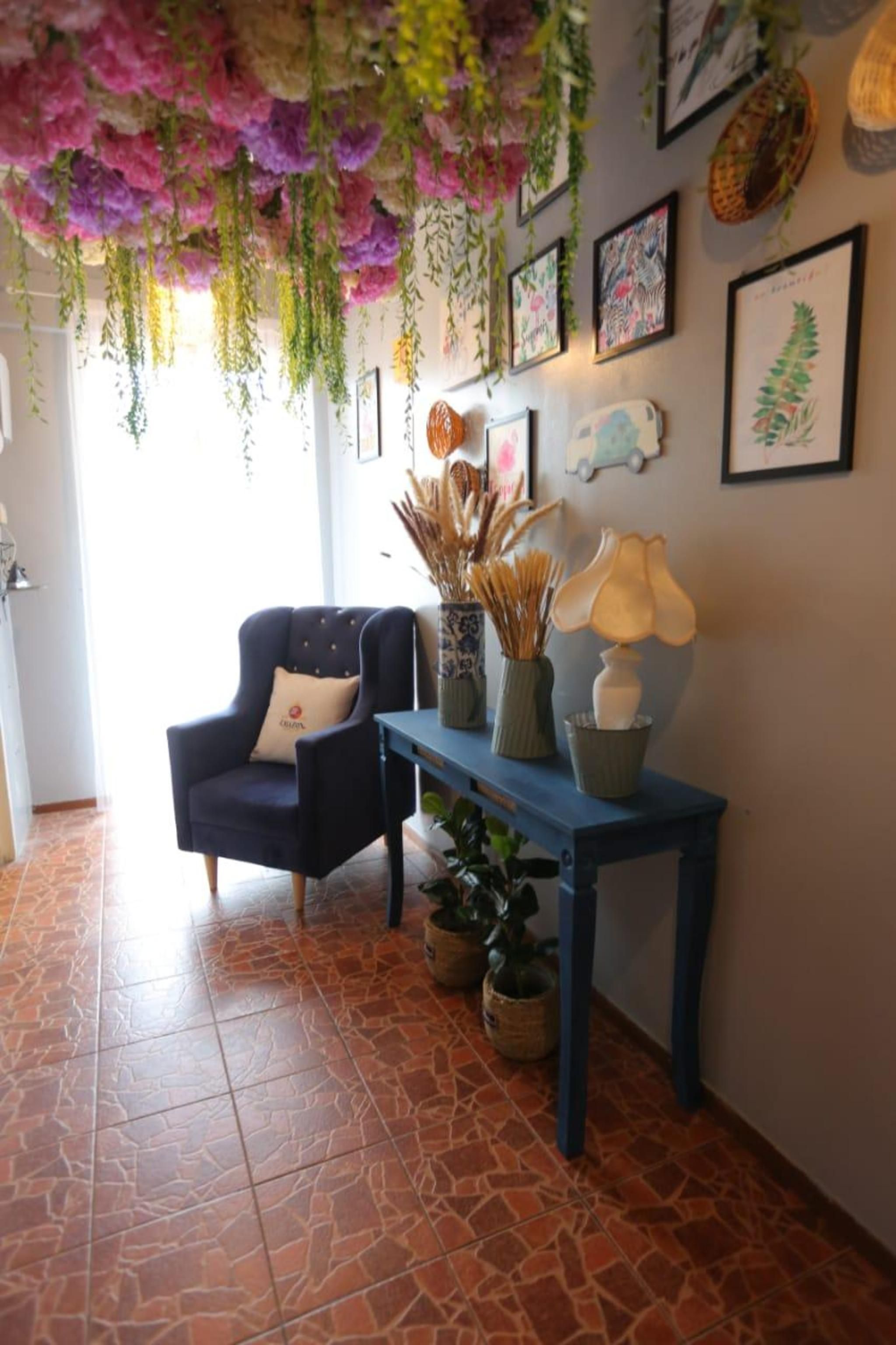 A room with a blue armchair, a blue table with decorative items, hanging plants, framed artwork, and a tiled floor.