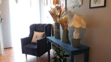 A room with a blue armchair, a blue table with decorative items, hanging plants, framed artwork, and a tiled floor.