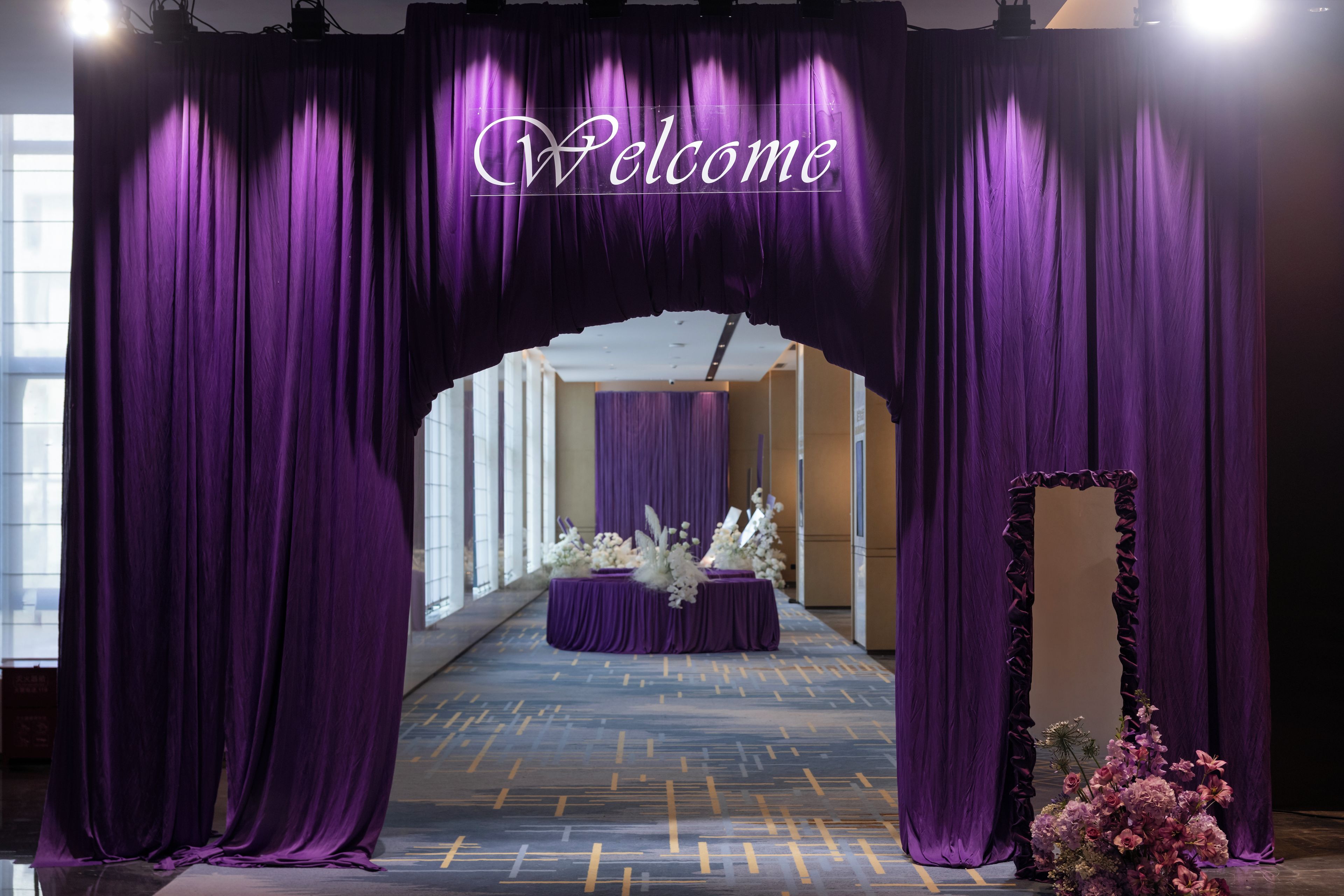 A purple curtain arch with 'Welcome' written on it, leading to a reception area with a floral arrangement.