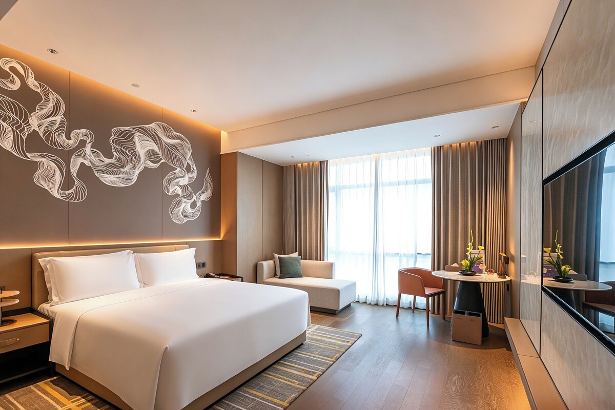 Una habitación de hotel moderna con una cama grande, un sofá, un comedor y un amplio ventanal con cortinas.
