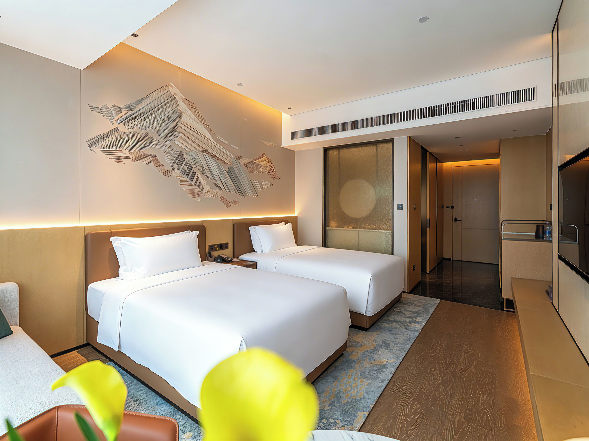 A hotel room with two beds, a large abstract wall art piece, a bathroom with a mirror, and a hallway leading to another room.