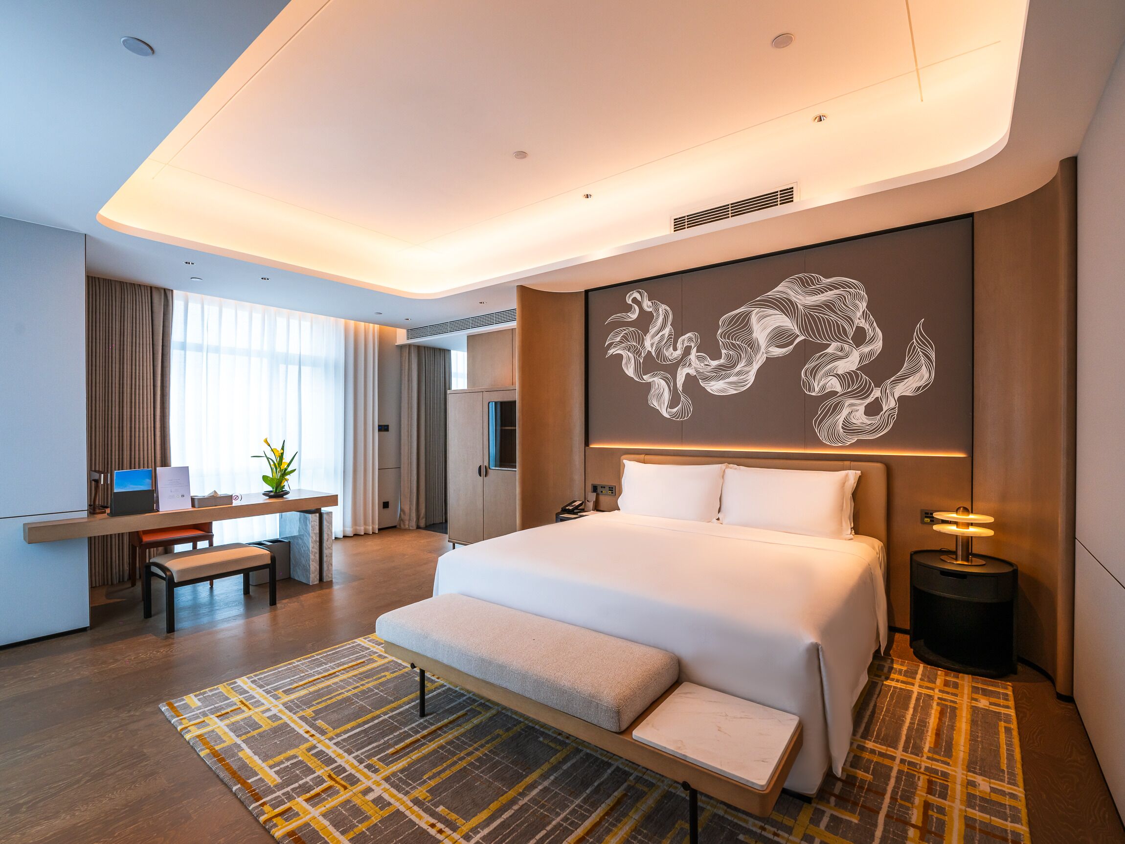 A modern hotel room with a large bed, a desk, a chair, a small table, a lamp, and a decorative wall piece.