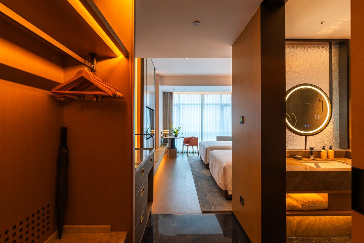 A hotel room with a bed, a desk, a chair, a bathroom with a mirror, and a view of the outside.