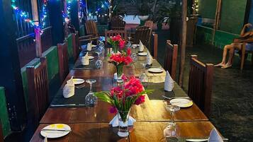 Una larga mesa de madera dispuesta para una comida, con sillas a ambos lados, bajo un espacio cubierto con techo de madera y luces colgantes.
