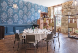 A dining area with a round table set for six, surrounded by chairs, with a patterned wallpaper and a stone wall in the background. - Hotel-spa L'essenciel - Château Des Pères (Piré-Chancé)