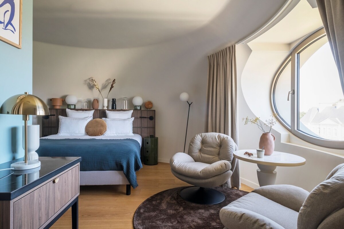 A bedroom with a bed, bedside table, chair, and a round window.