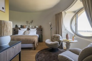 A modern hotel room with a bed, a round table with a vase and a bottle, a chair, and a view through a large window. - Hotel-spa L'essenciel - Château Des Pères (Piré-Chancé)
