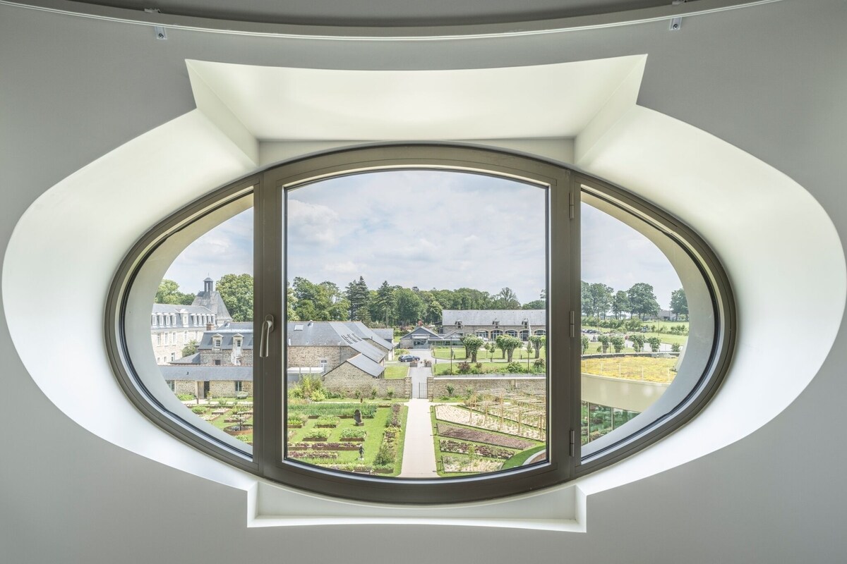 Habitació amb un gran ull de bou circular que dona vistes a un jardí i edificis.