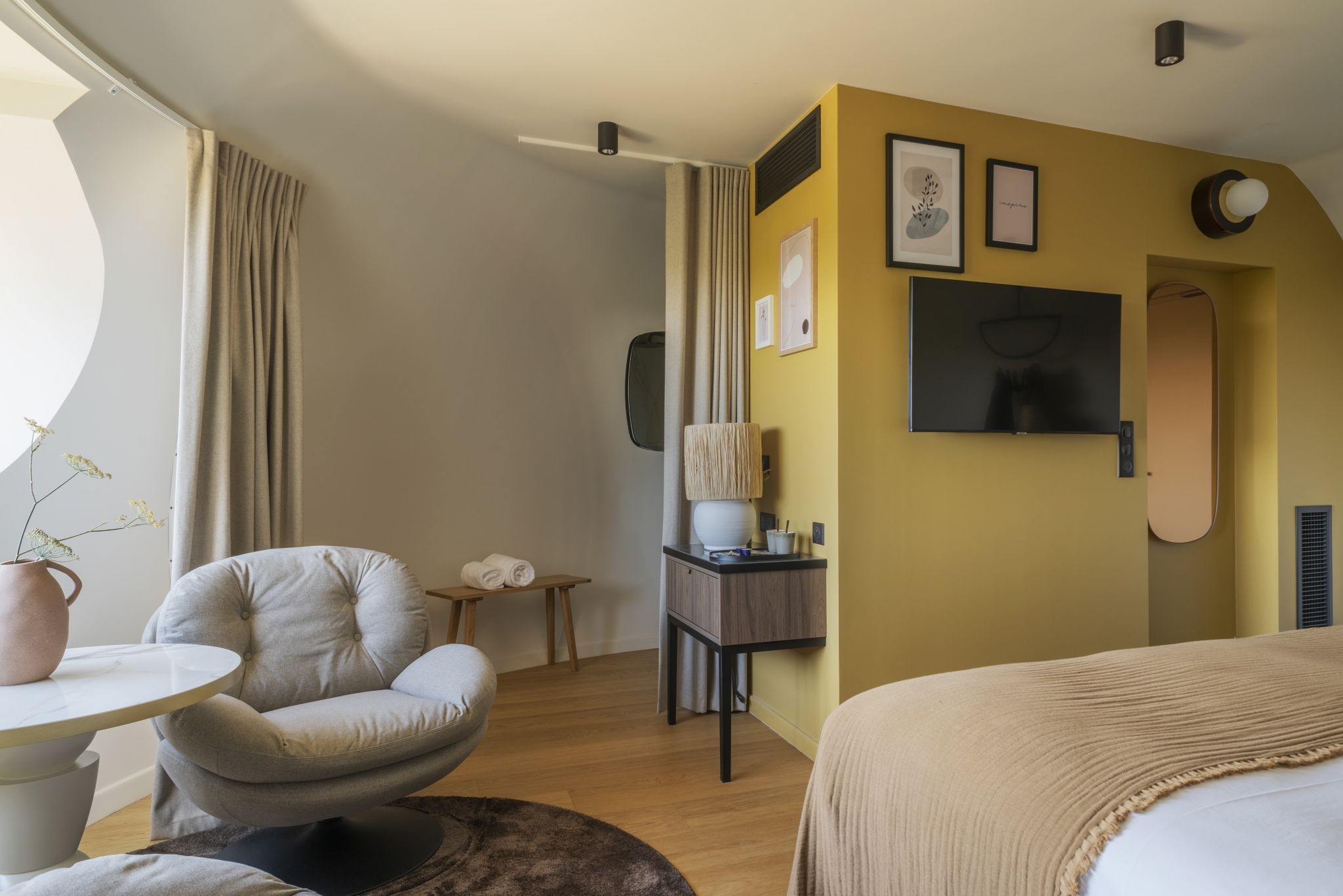A hotel room with a bed, a round table, a chair, a small table with a vase, and a wall-mounted TV.