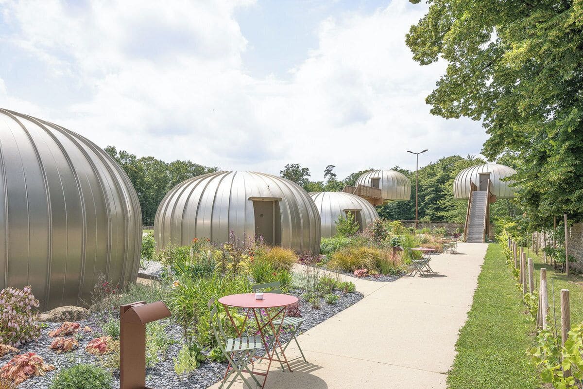 Un sendero conduce a estructuras esféricas con puertas, rodeado de un jardín con diversas plantas y una pequeña mesa con sillas.