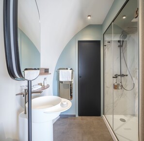 A modern bathroom with a curved mirror, white sink, and a glass-enclosed shower. - Hotel-spa L'essenciel - Château Des Pères (Piré-Chancé)