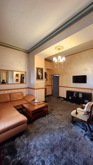 A living room with a leather sofa, a wooden coffee table, a patterned rug, a chandelier, a television, and a framed picture on the wall. - Nf Palace Old City Bucharest (Bucharest)