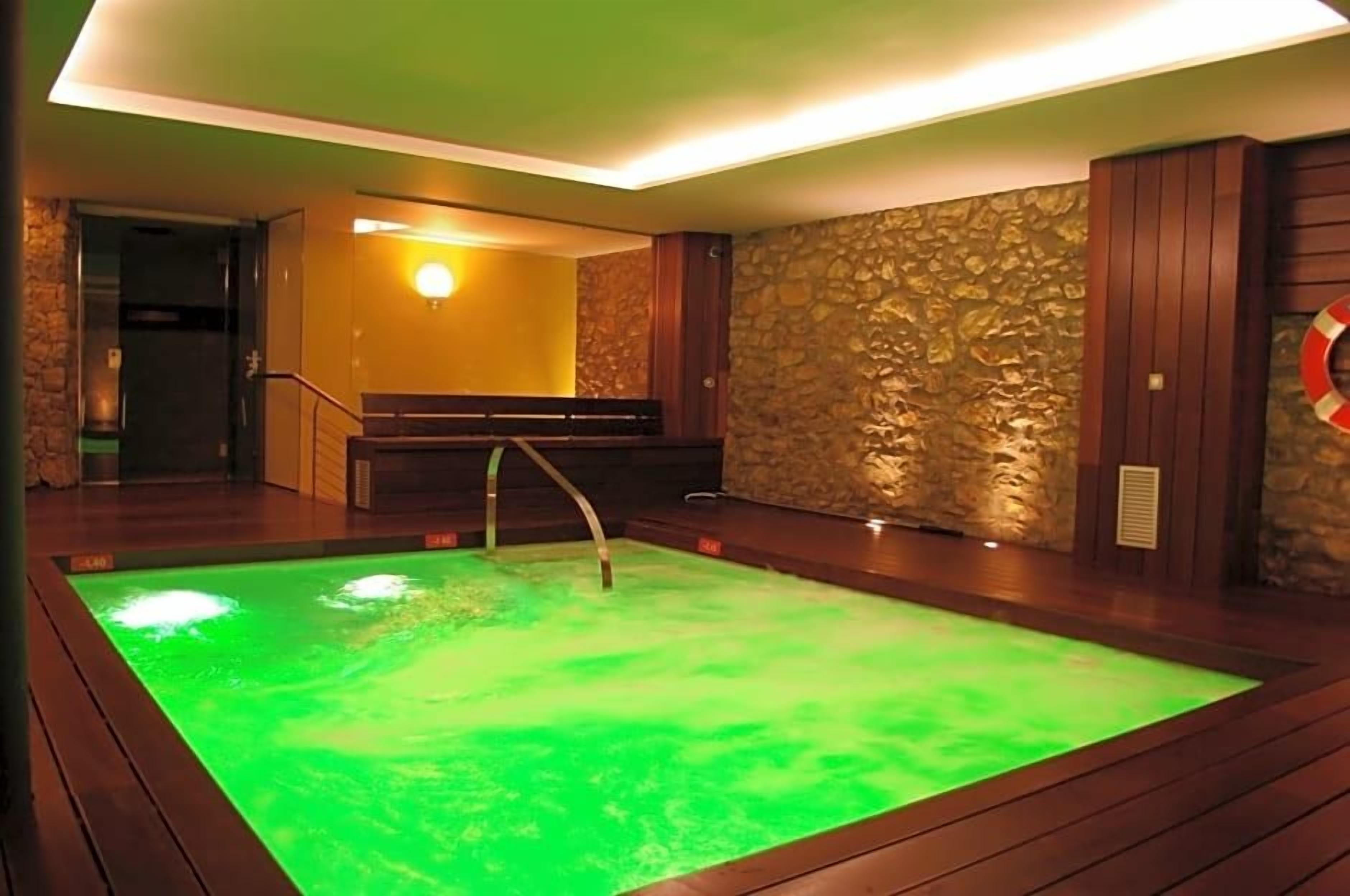 A spa area with a heated pool, wooden flooring, stone wall, and a bench.