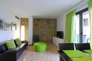 A living room with a stone wall, a sofa, a green ottoman, a TV, and a dining area with chairs. - Dolcet L Hotel (Alàs i Cerc)