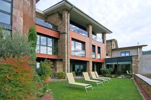 Een modern gebouw met een gevel van natuursteen, grote ramen en een verzorgd gazon met loungestoelen.