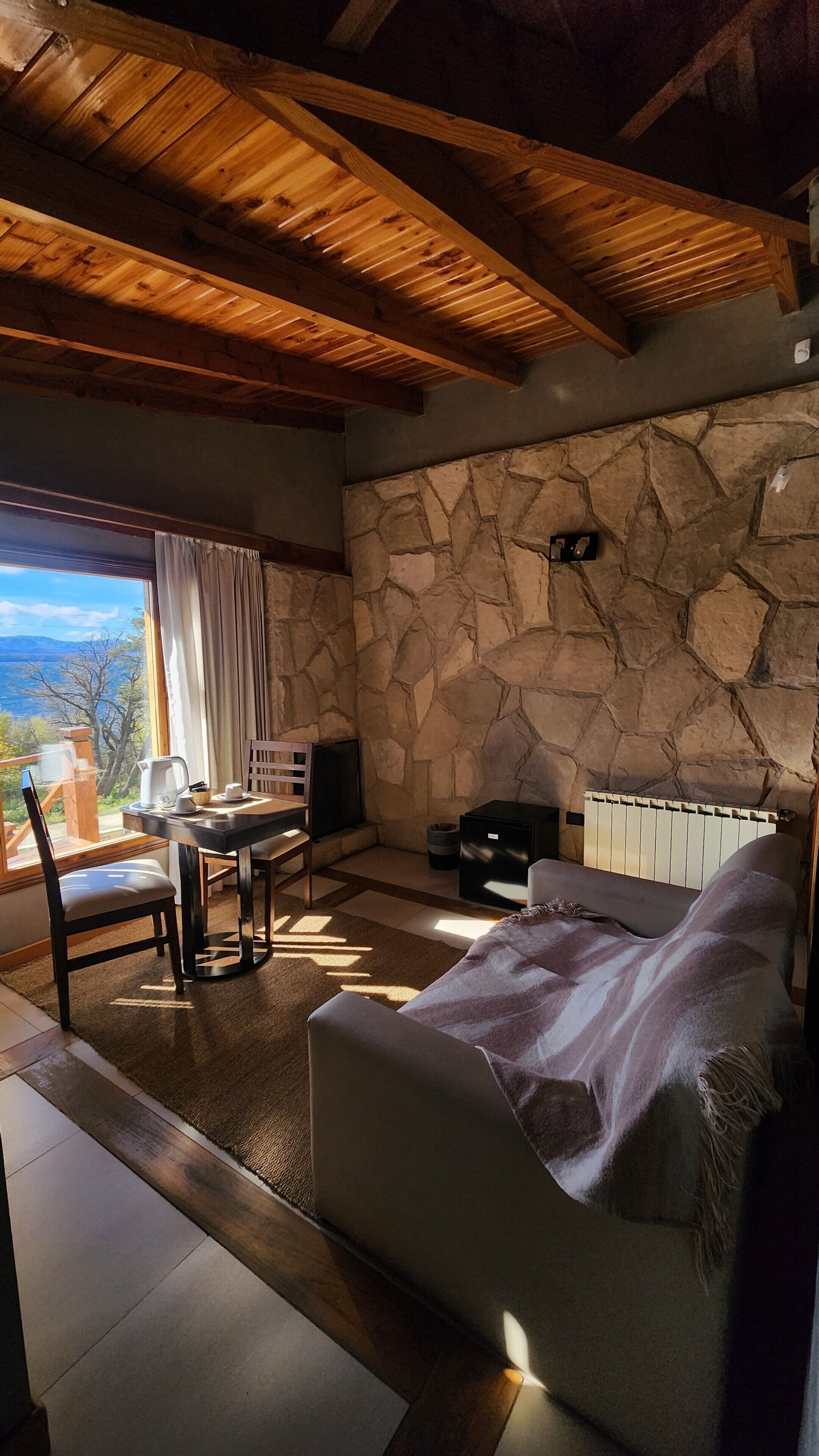 Un cuarto acogedor con pared de piedra, vigas de madera en el techo, chimenea, sofá, mesa de comedor con sillas y vista al exterior.