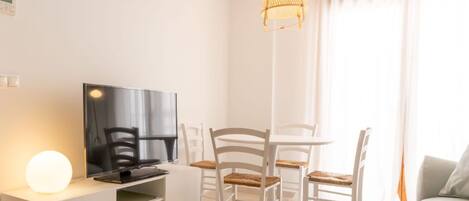 A modern living room with a flat-screen TV, a white entertainment unit, a dining table with chairs, and a hanging light fixture.