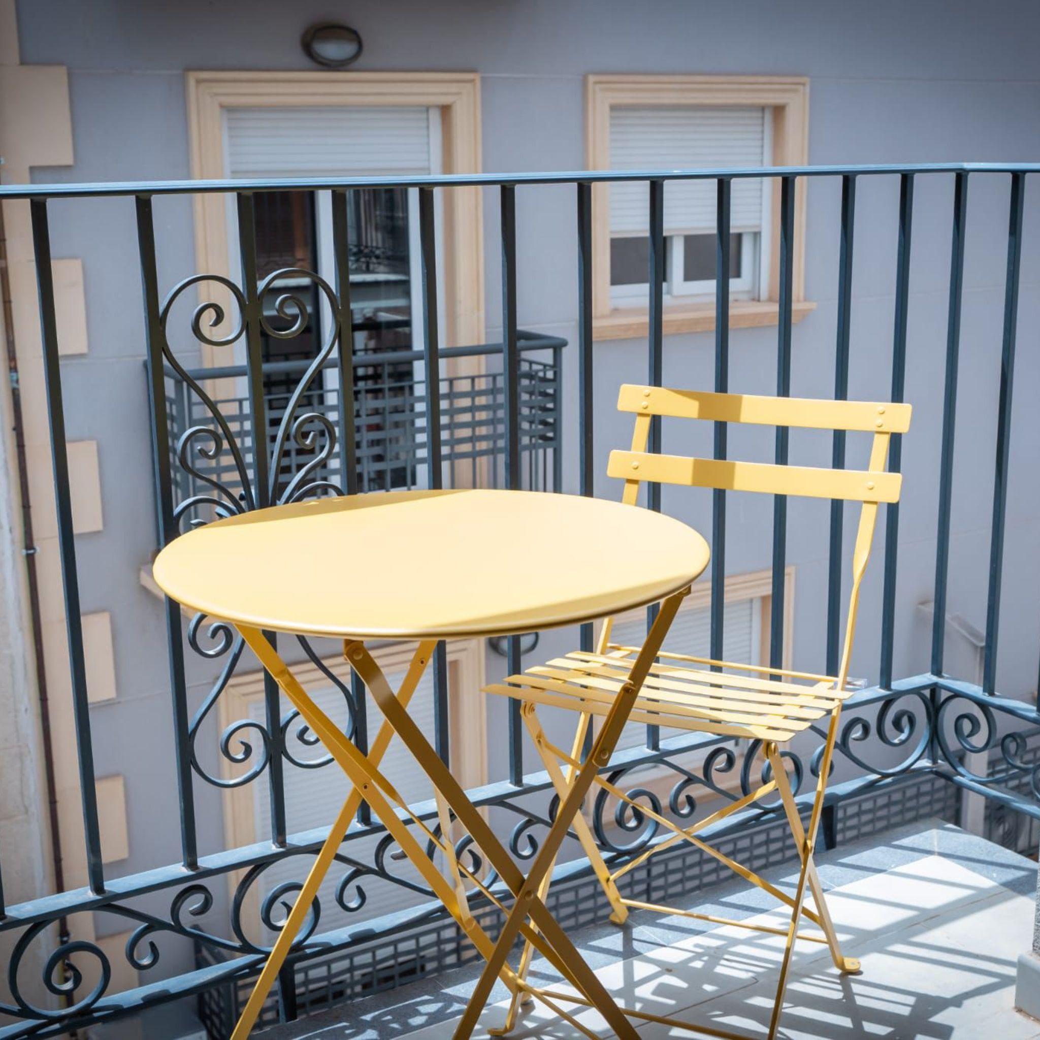 Un balcon avec une table et une chaise jaunes, des rampes en métal et une fenêtre avec des stores.