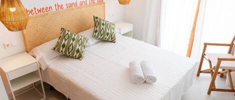 Una cama bien hecha con almohadas, una mesita de noche, una lámpara y una silla en una habitación luminosa con un cartel en la pared.