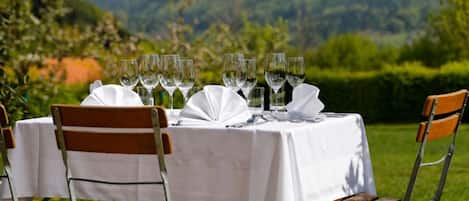 An outdoor dining setup with a table set for four, surrounded by chairs, in a lush green landscape with mountains in the background.