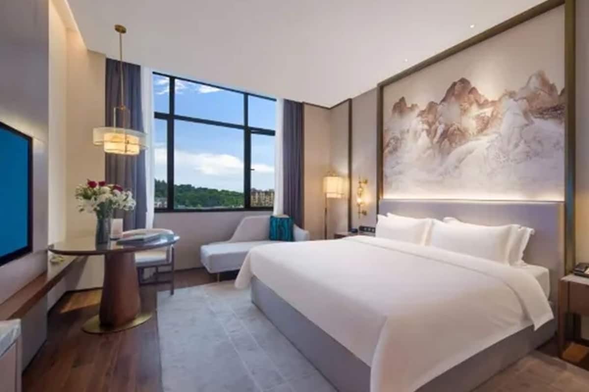 Una habitación de hotel moderna con una cama grande, un escritorio con silla, un televisor y un amplio ventanal con vistas a la montaña.