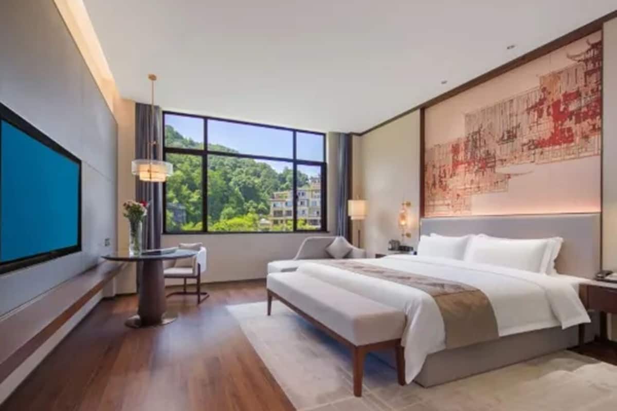 Habitación de hotel moderna con una cama grande, un escritorio, una silla y un amplio ventanal con vistas a zonas verdes y edificios.
