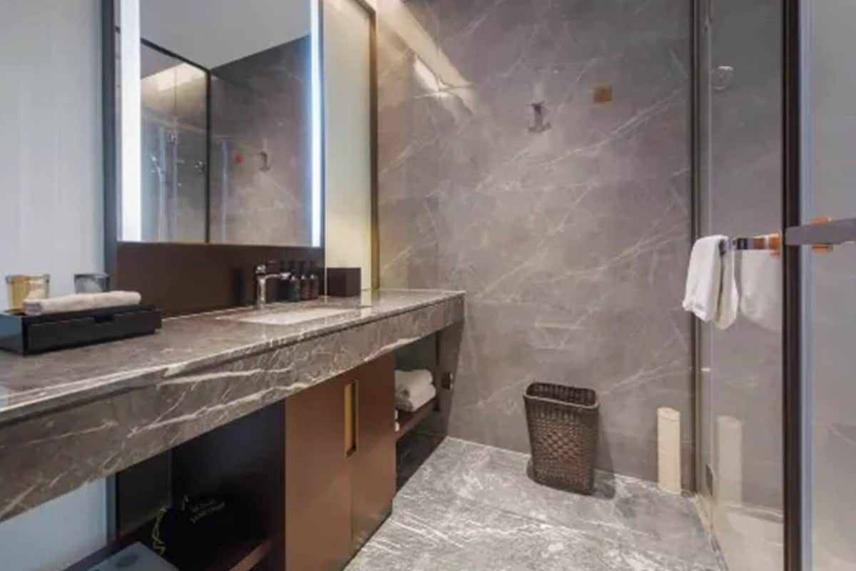 A modern bathroom with a marble countertop, a large mirror, a glass shower enclosure, and a small trash bin.