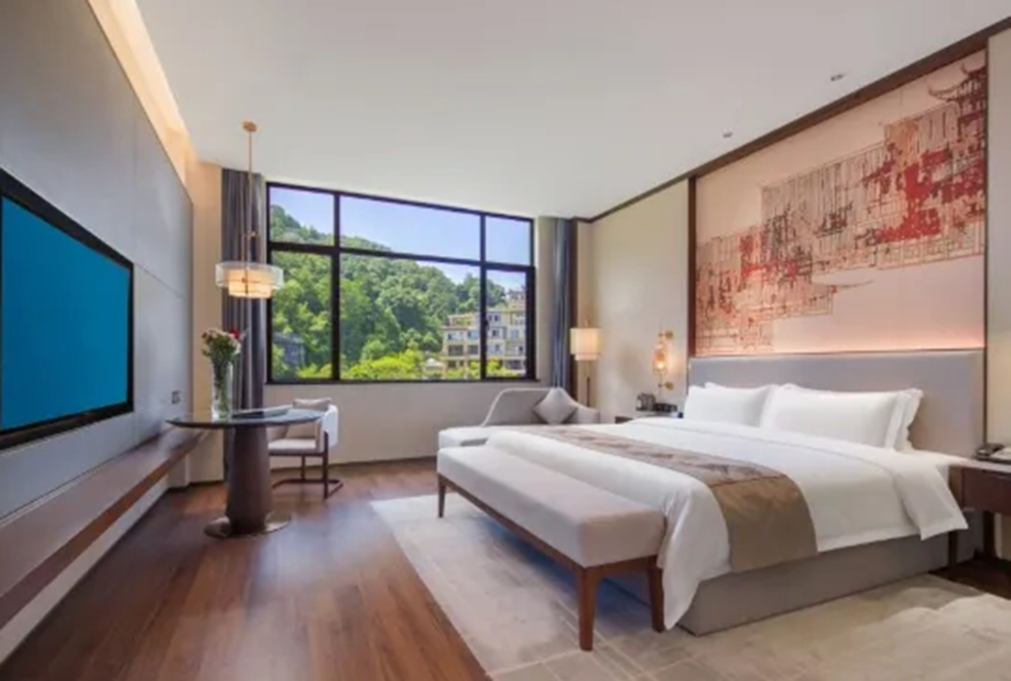 A modern hotel room with a large bed, a desk, a chair, and a large window offering a view of greenery and buildings.