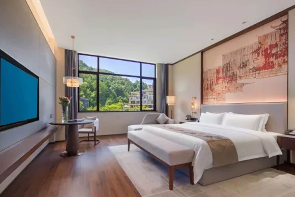 Habitación de hotel moderna con una cama grande, un escritorio, una silla y un amplio ventanal con vistas a zonas verdes y edificios.