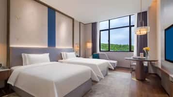 A hotel room with two beds, a desk, a chair, a round table with a vase, and a large window offering a view of greenery.