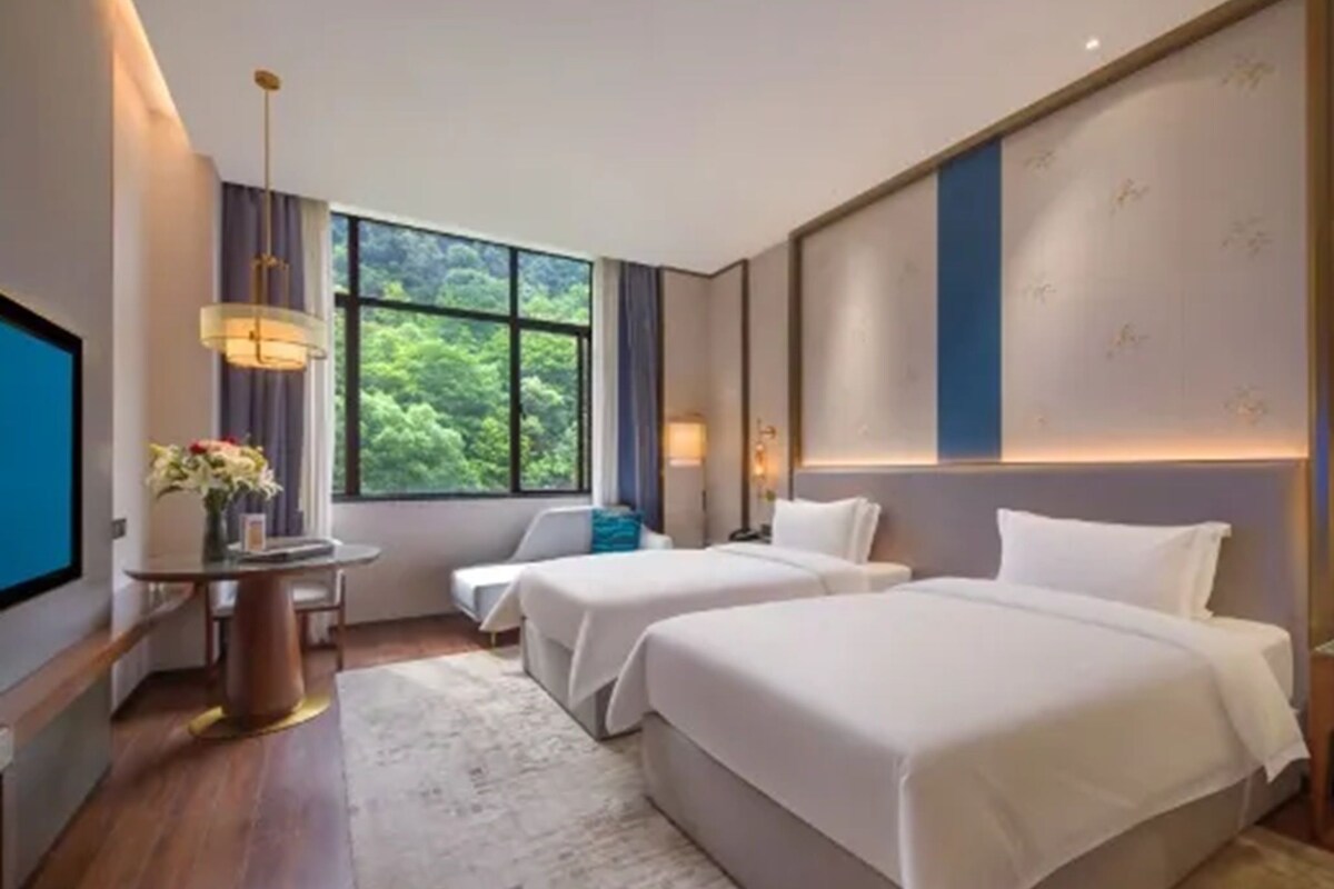 Habitación de hotel con dos camas, un escritorio con lámpara, televisión y un ventanal con vistas al verdor.