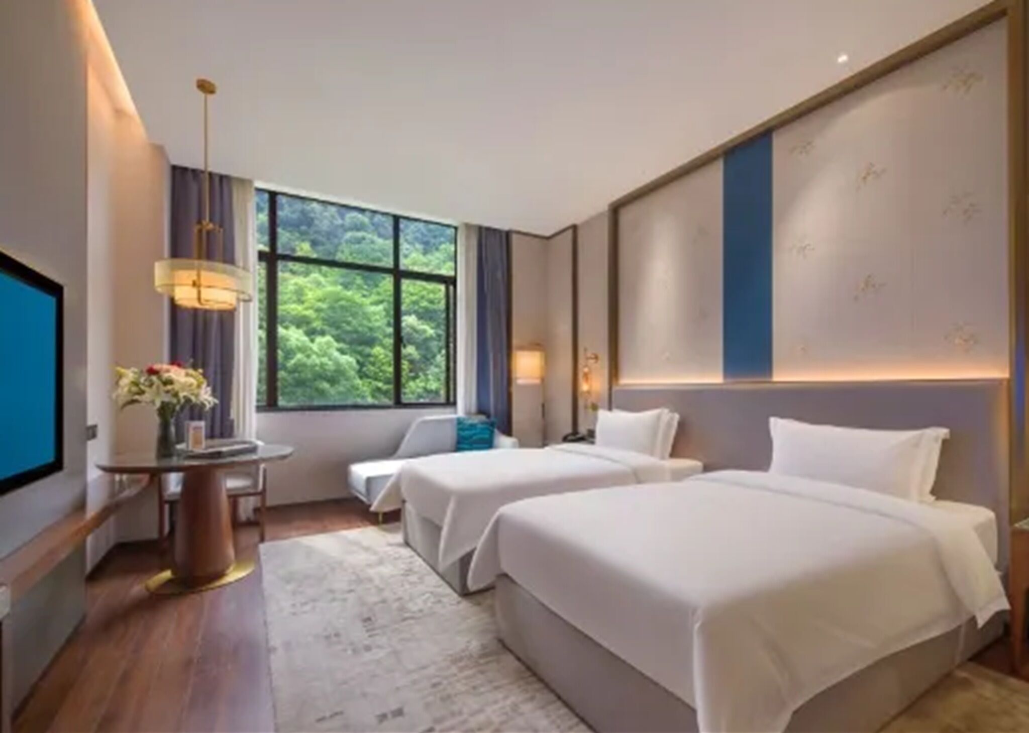 Une chambre d’hôtel avec deux lits, un bureau avec une lampe, une télévision et une grande fenêtre donnant sur un paysage verdoyant.