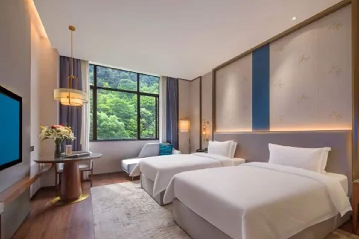 Habitación de hotel con dos camas, un escritorio con lámpara, televisión y un ventanal con vistas al verdor.