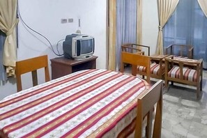 Una habitación con una cama, un televisor sobre un soporte de madera y dos sillas de madera con cojines.