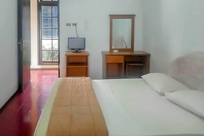 Habitación de hotel con cama, televisor sobre un atril de madera, espejo sobre un escritorio y puerta que da a un espacio exterior.