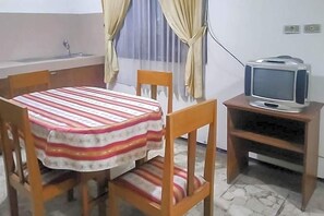 A small room with a dining table, chairs, a television on a wooden stand, a sink, and a window with curtains. - Mataram Cottage Syariah Redpartner (Cipanas)