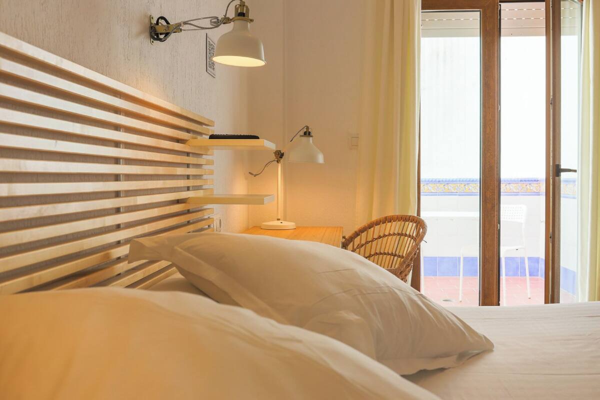 A hotel room with a bed, two bedside lamps, a wicker chair, and a view of the outside.
