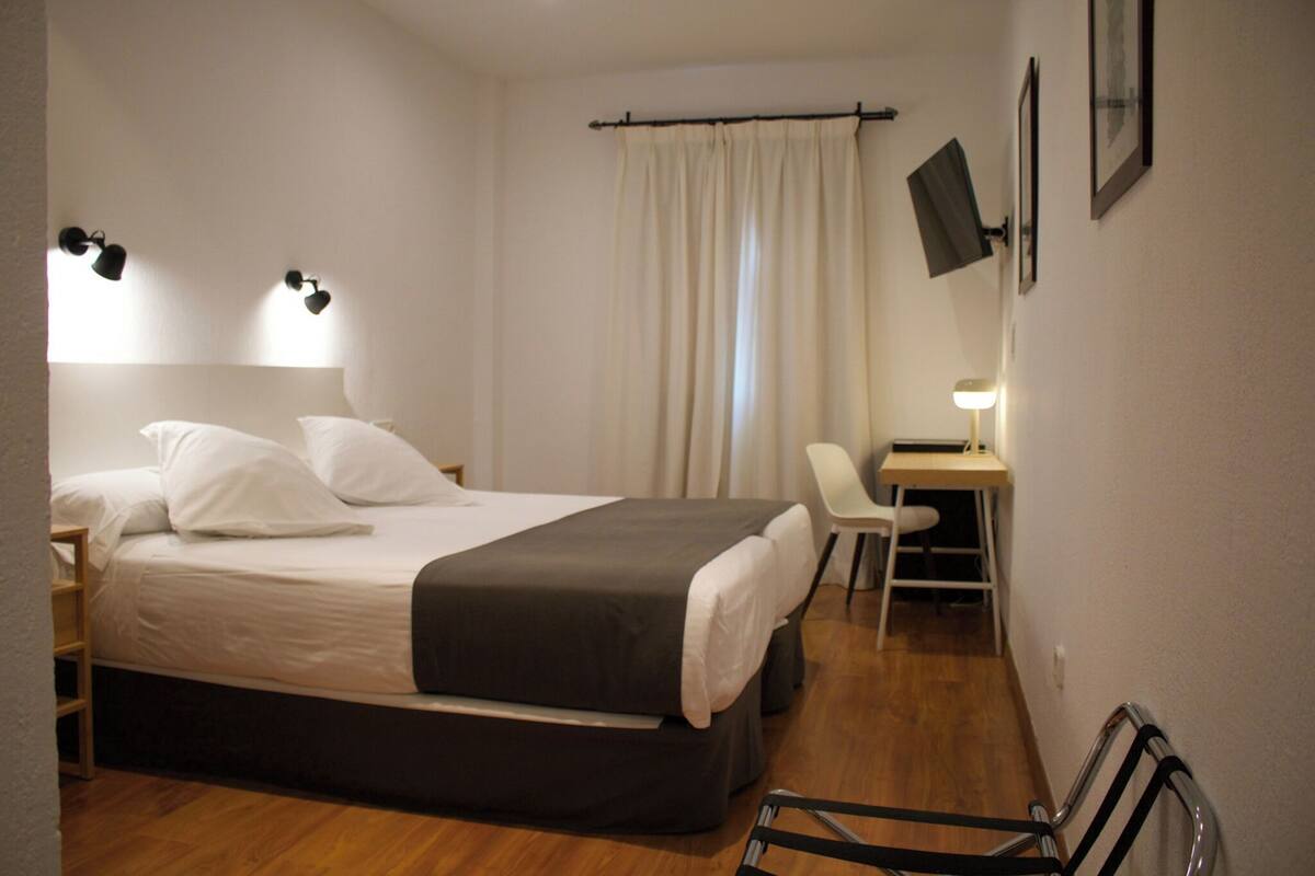 A bedroom with a bed, bedside tables, a desk, a chair, a TV, and a curtain.