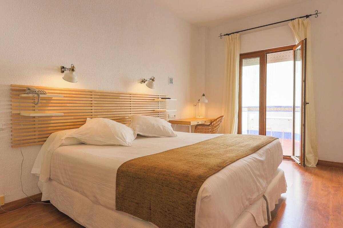 A bedroom with a bed, bedside lamps, a wooden headboard, a desk with a chair, and a balcony with curtains.
