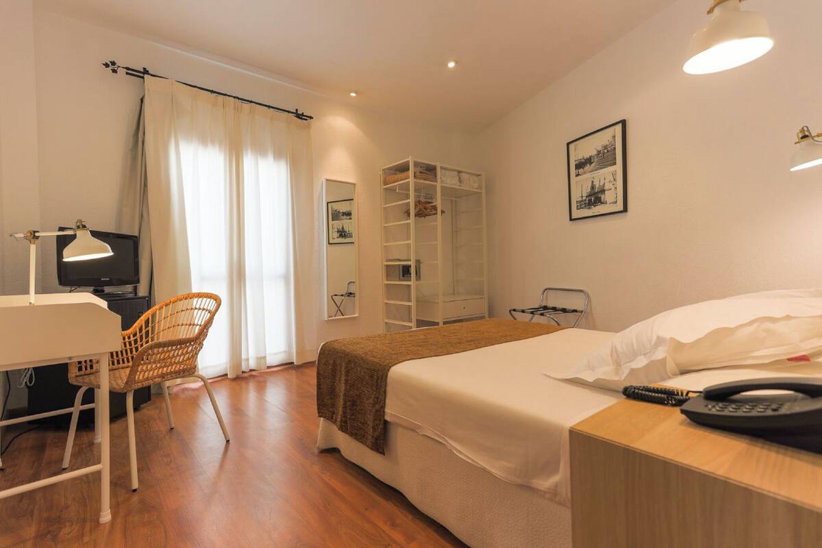 A hotel room with a bed, a desk with a lamp, a chair, a wardrobe, and a telephone.