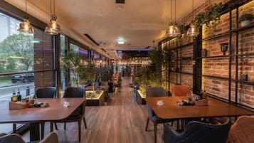 A restaurant interior with wooden tables and chairs, hanging pendant lights, brick walls, and potted plants.
