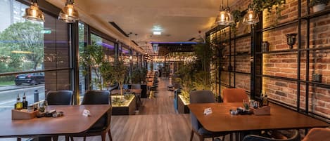 Interior de restaurante con mesas y sillas de madera, lámparas colgantes, paredes de ladrillo y plantas en macetas.