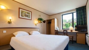 Et hotelværelse med seng, skrivebord, fjernsyn og et vindue med gardiner.