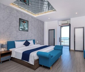 Habitación de hotel moderna con cama, sofá azul, mesita de noche, un cuadro, una lámpara en el techo y una puerta que da a un balcón.