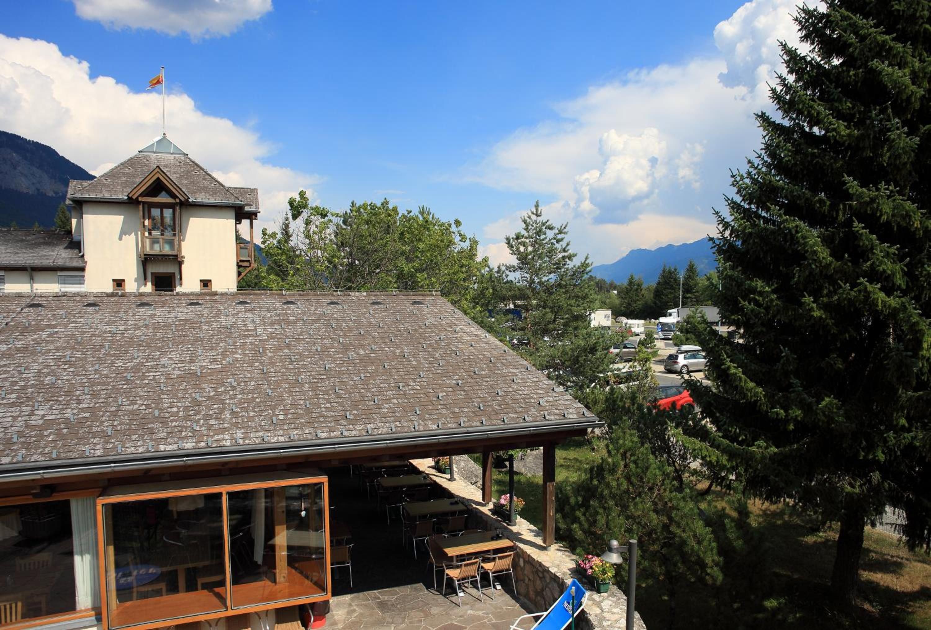 A building with a sloped roof, a covered outdoor seating area with tables and chairs, and a mountain backdrop.