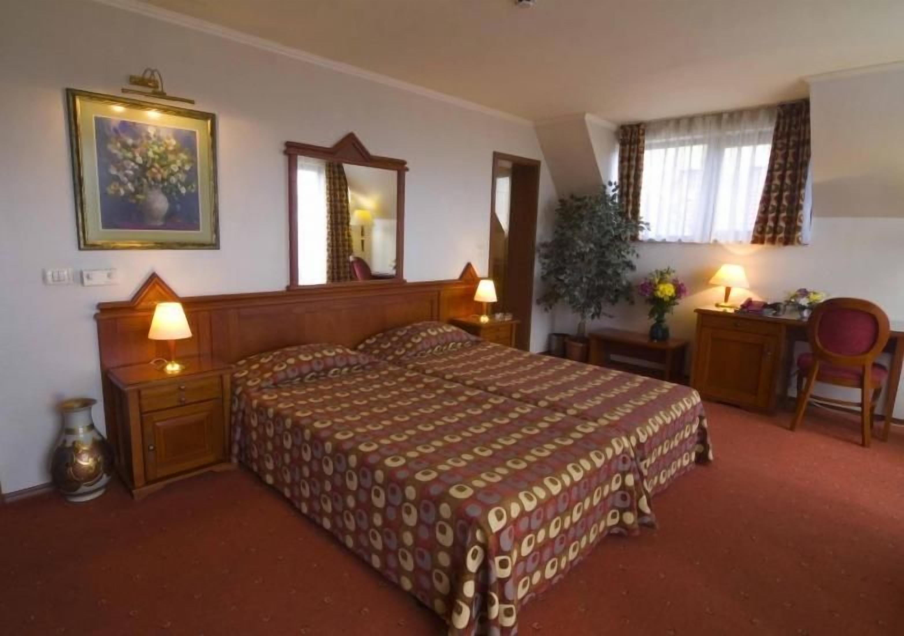 A bedroom with a bed, bedside tables, a desk, a chair, a mirror, and a potted plant.