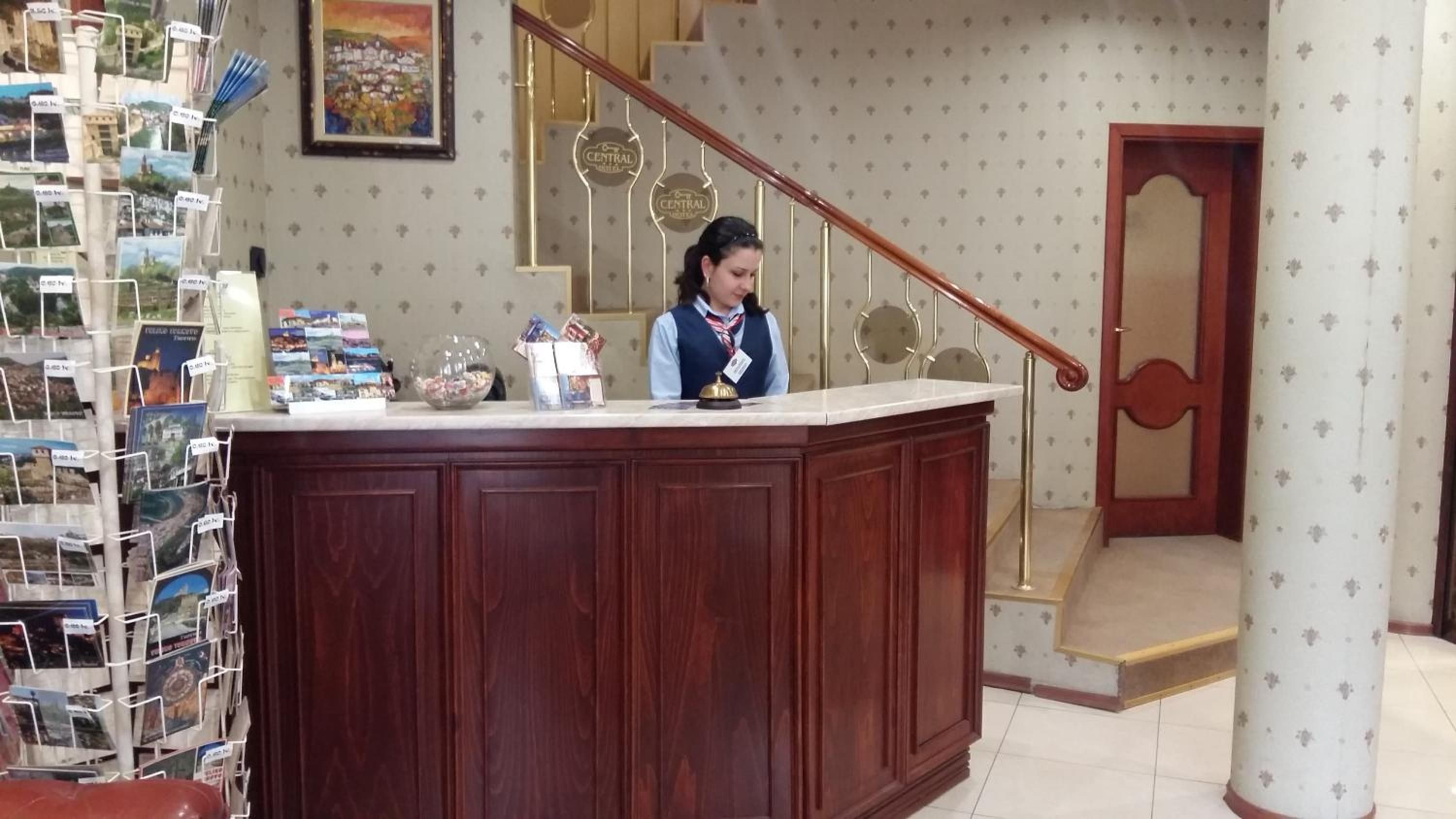 Una recepción con un mostrador de madera, un timbre y una placa de identificación. Se ve una escalera con barandilla y una puerta al fondo.