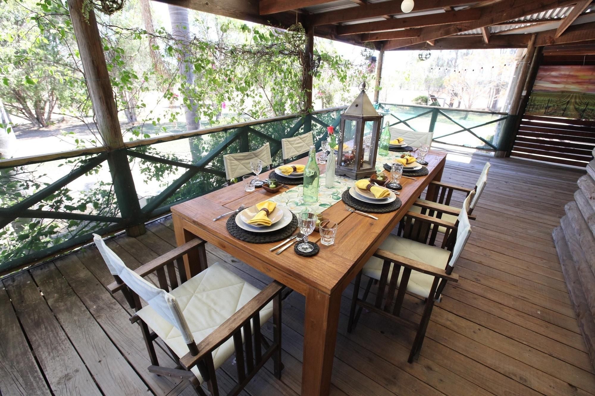 A wooden deck with a table set for dining, surrounded by chairs, and a view of greenery through a wooden railing.