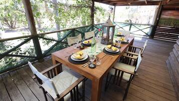 A wooden deck with a table set for dining, surrounded by chairs, and a view of greenery through a wooden railing.